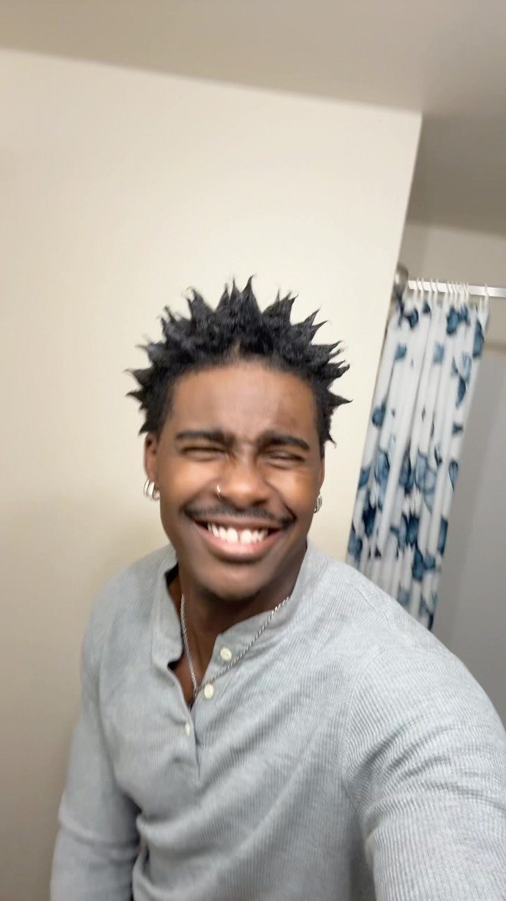 Smiling Black man with kinky hair styled into sharp spikes wearing a grey shirt.