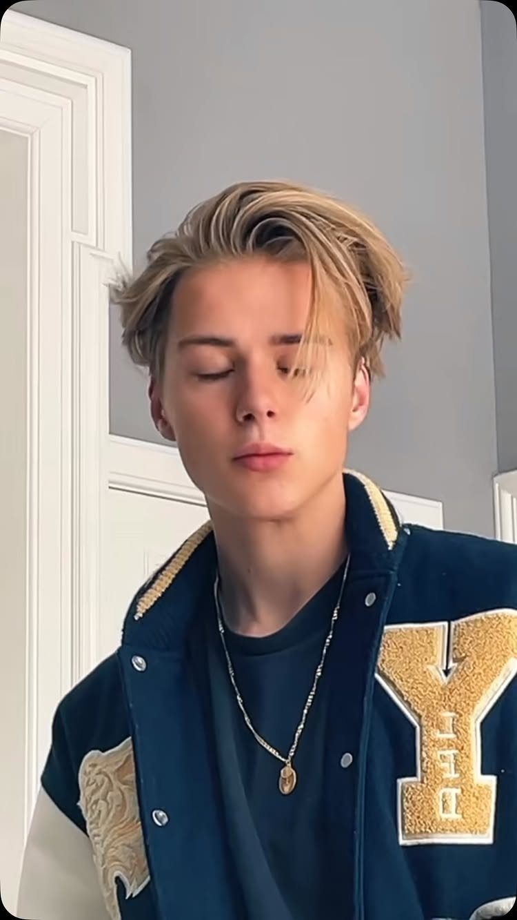 Young man with medium-length blonde hair swept back wearing a navy varsity jacket.