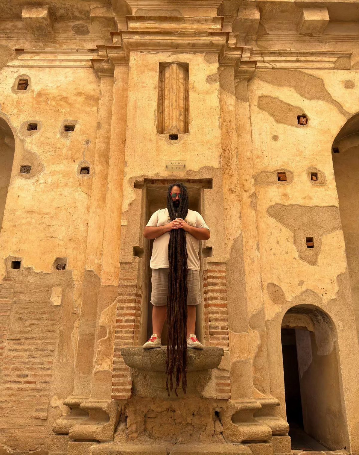 Man with extremely long dark brown dreadlocks standing before an ancient stone wall.