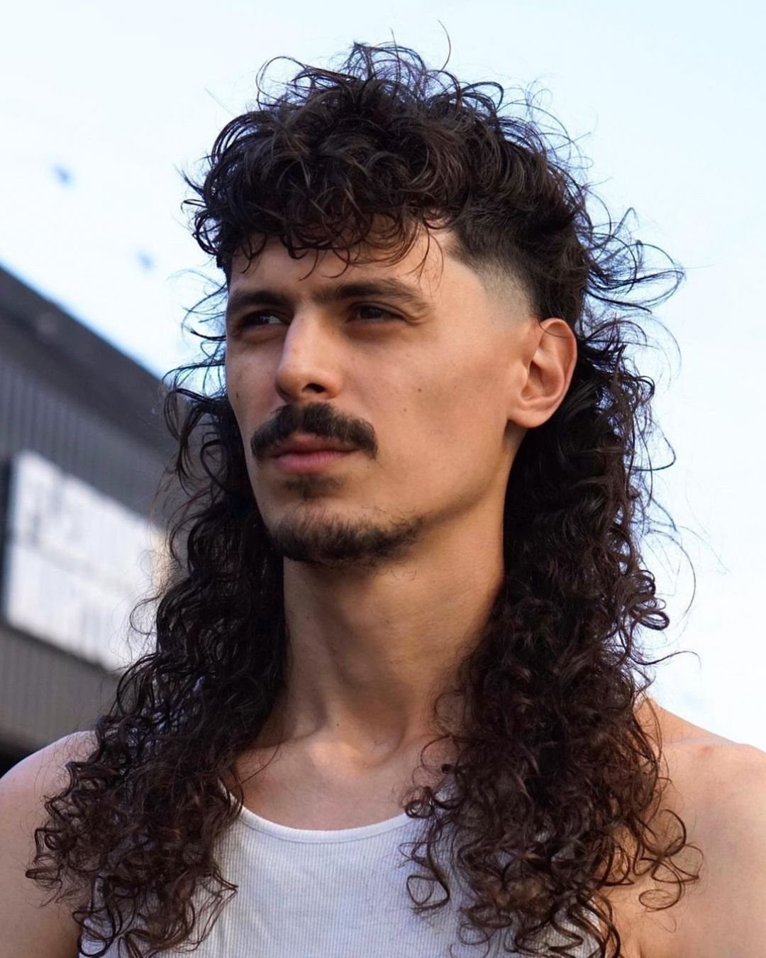 Man with a long curly mullet hairstyle and a mustache wearing a white tank top.