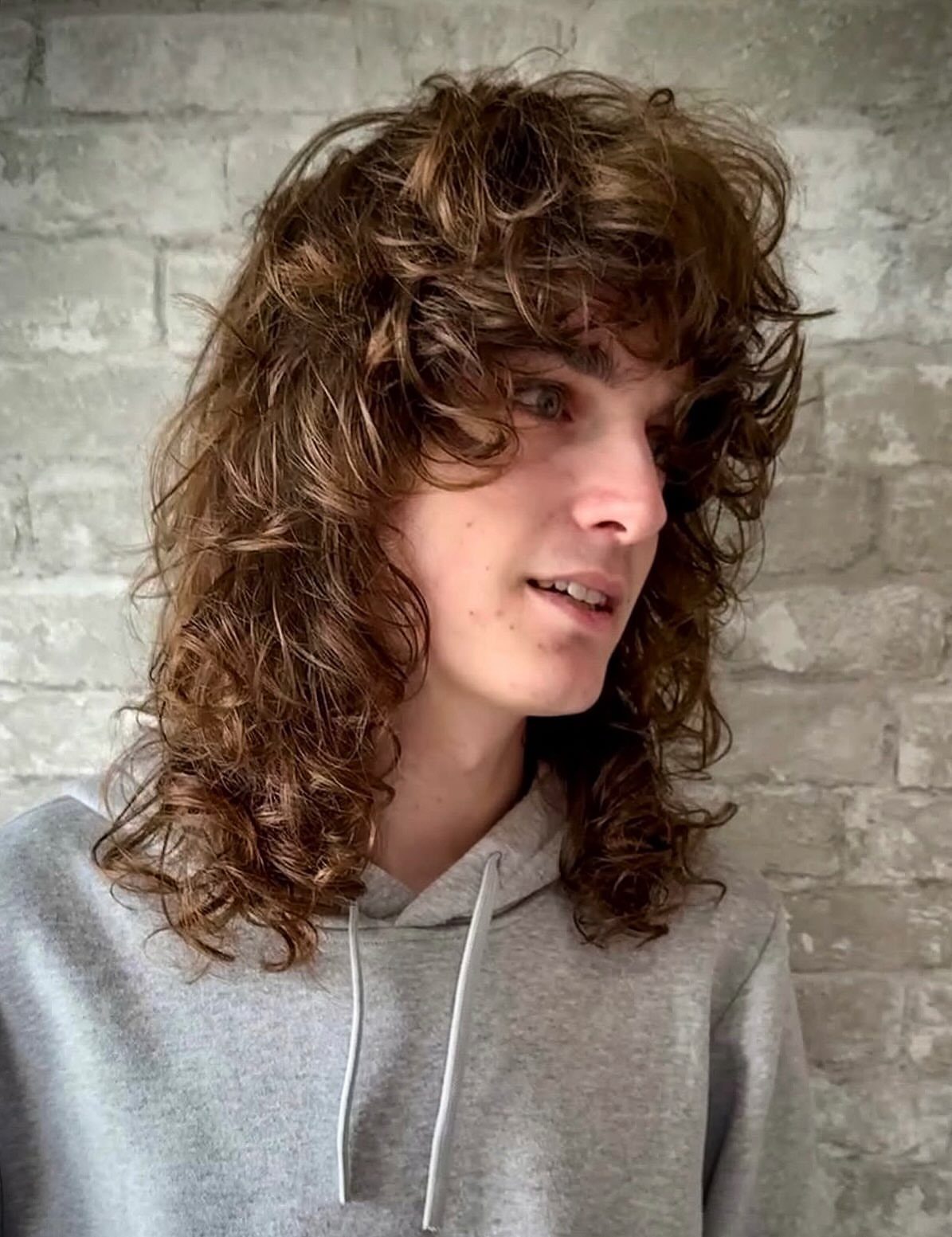 Young man with long brown curly shag haircut and fringe wearing grey hoodie.