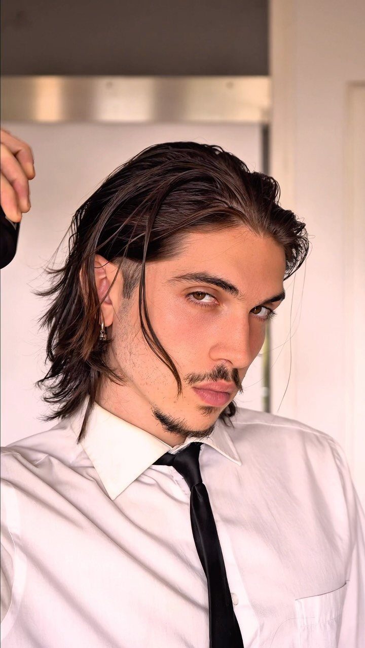 Man with long dark hair slicked back wearing a white shirt and black tie.