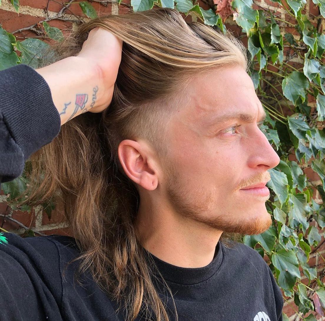 Man with long light brown hair and shaved sides posing against ivy wall.