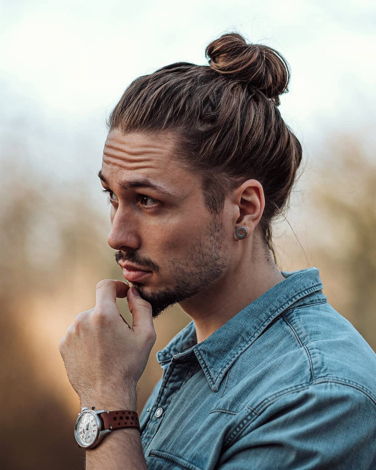 Man with brown hair tied in a high man bun wearing a denim shirt.