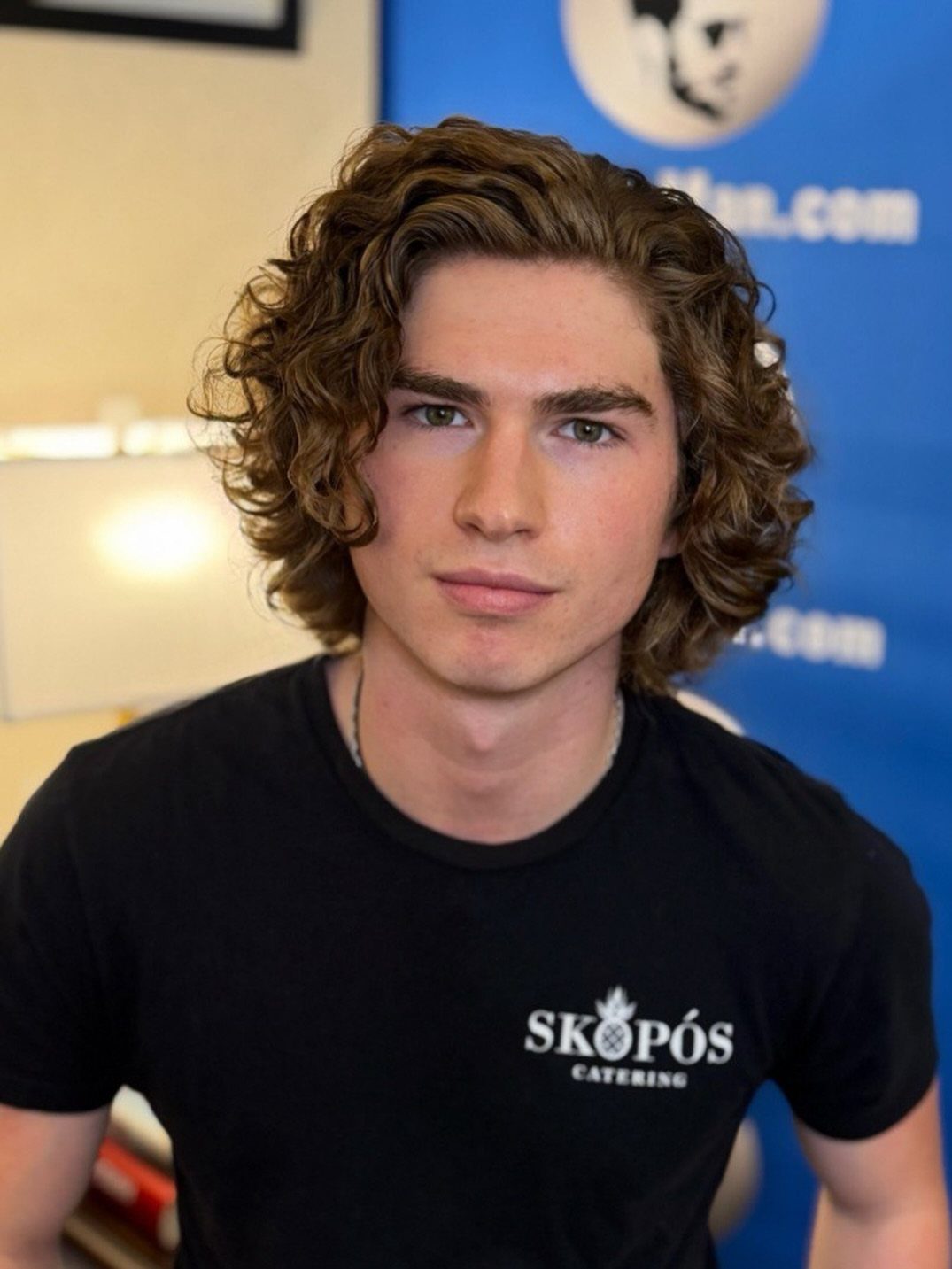 Young man with curly brown shoulder-length hair wearing a black t-shirt.