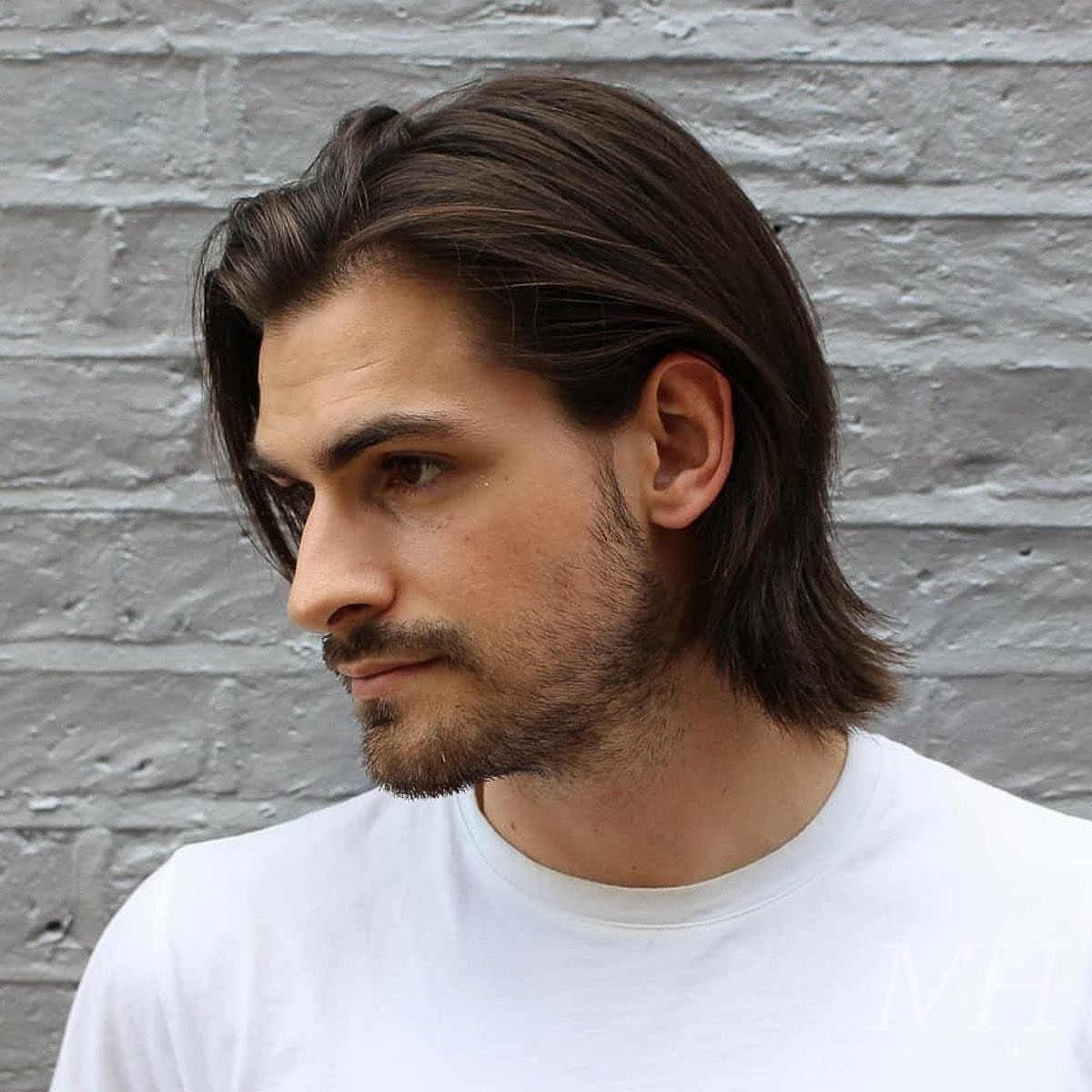 Man with brown shoulder-length hair swept back and a side part against brick wall.