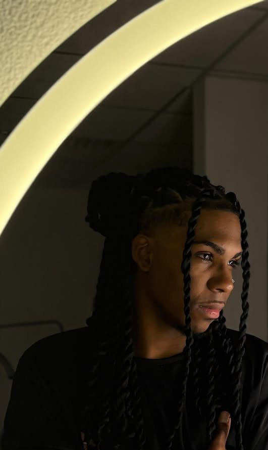 Young man with long black twisted dreadlocks posing near a curved white light.