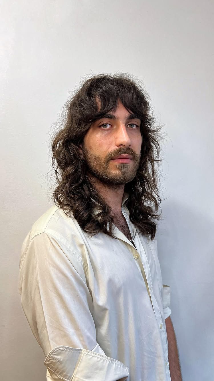 Man with long wavy brown hair and beard wearing a white shirt.