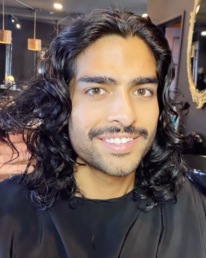 Man with shoulder-length wavy black hair and a mustache smiling in a salon.