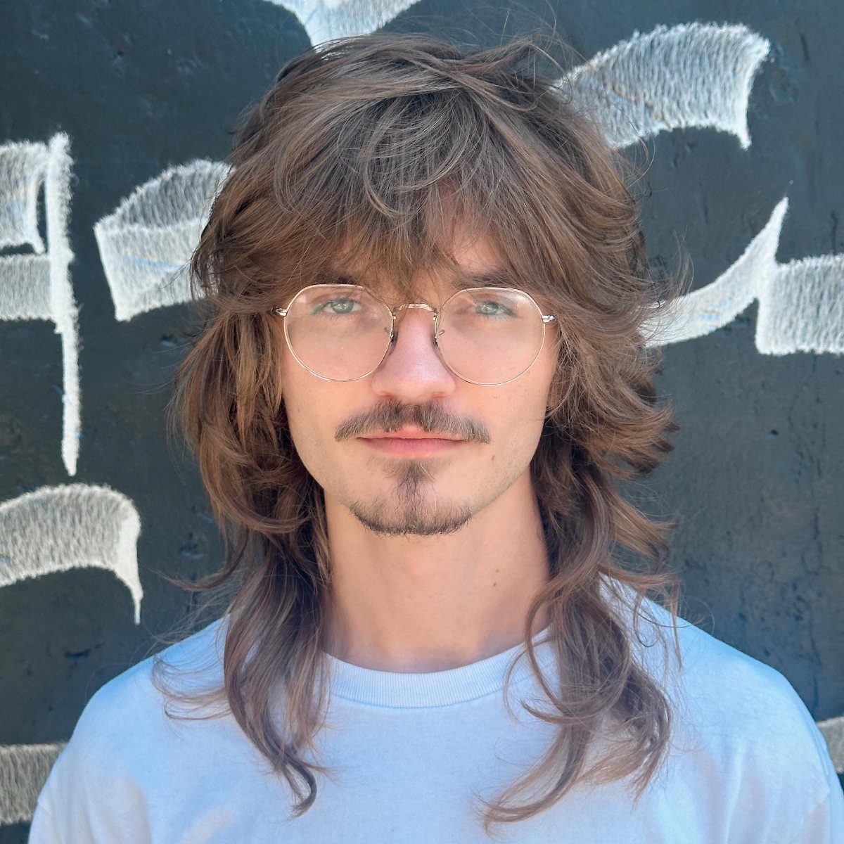 Man with a shaggy brown wolf cut hairstyle and round glasses.