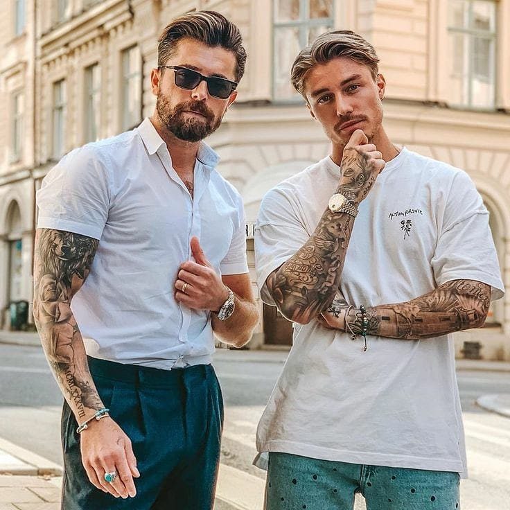 Two men with styled medium length hair and arm tattoos posing on a street.