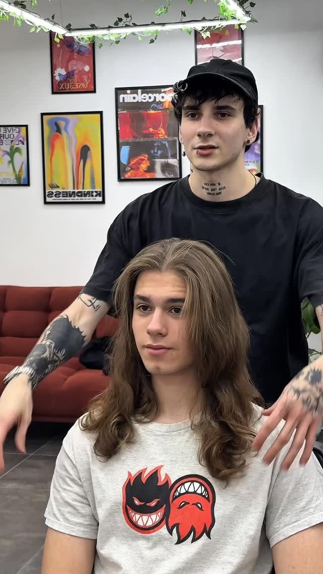 Man with long wavy brown hair posing in a hair salon with a stylist.