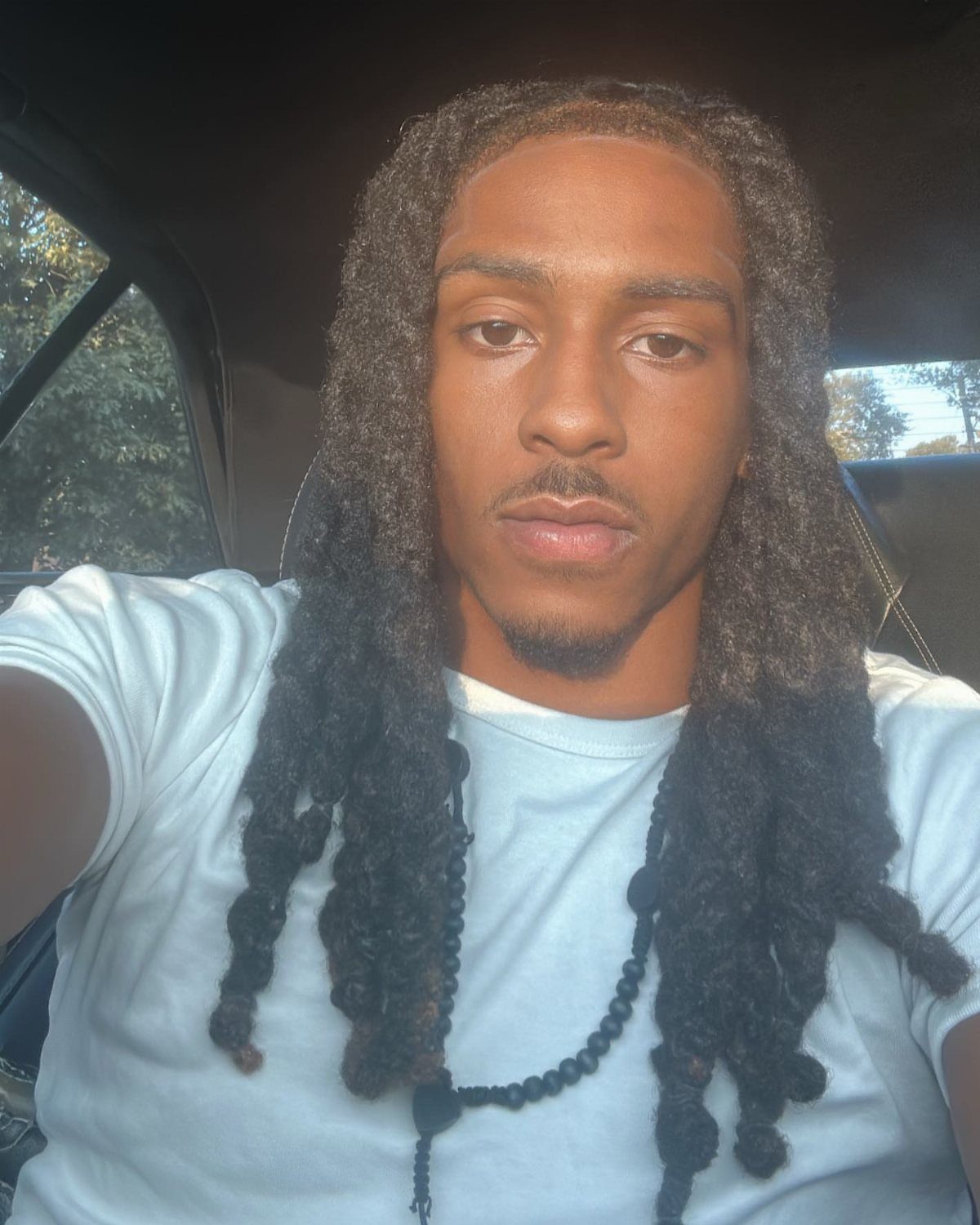 Man with mid-length brown locs wearing a white t-shirt and beaded necklace.
