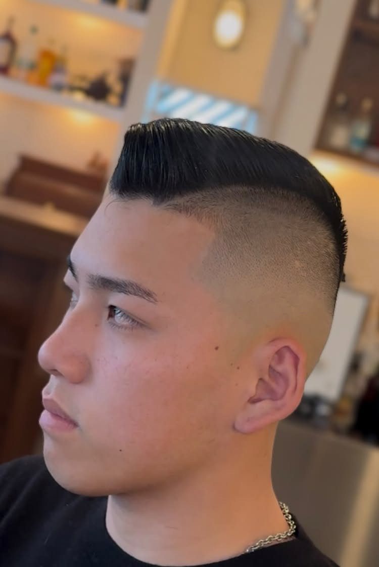 Man with a slicked-back hairstyle featuring a shaved hard part and mid skin fade.