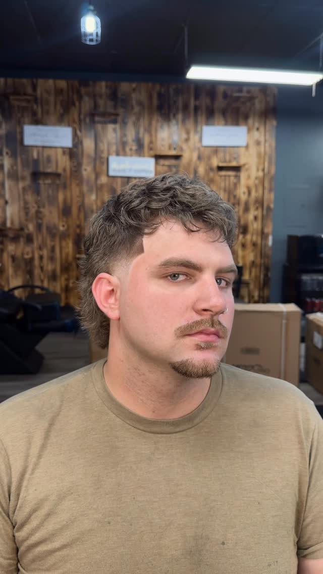 Man with a curly mullet haircut, mustache, and goatee in a barbershop setting.