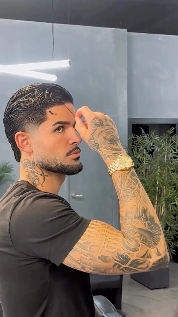 Man with slicked-back hair, groomed beard, and arm tattoos posing in a studio.