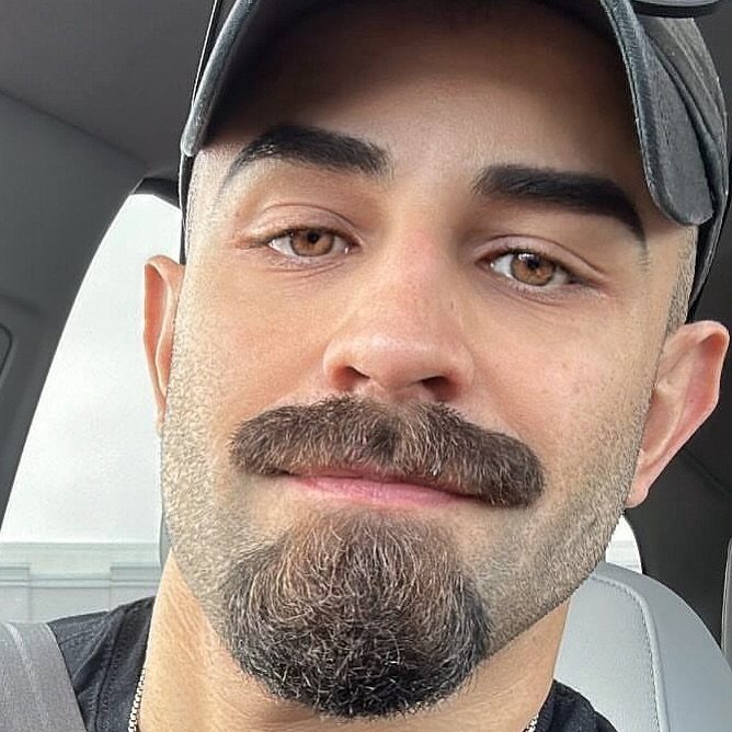 Man with a thick brown mustache and groomed goatee wearing a black cap.