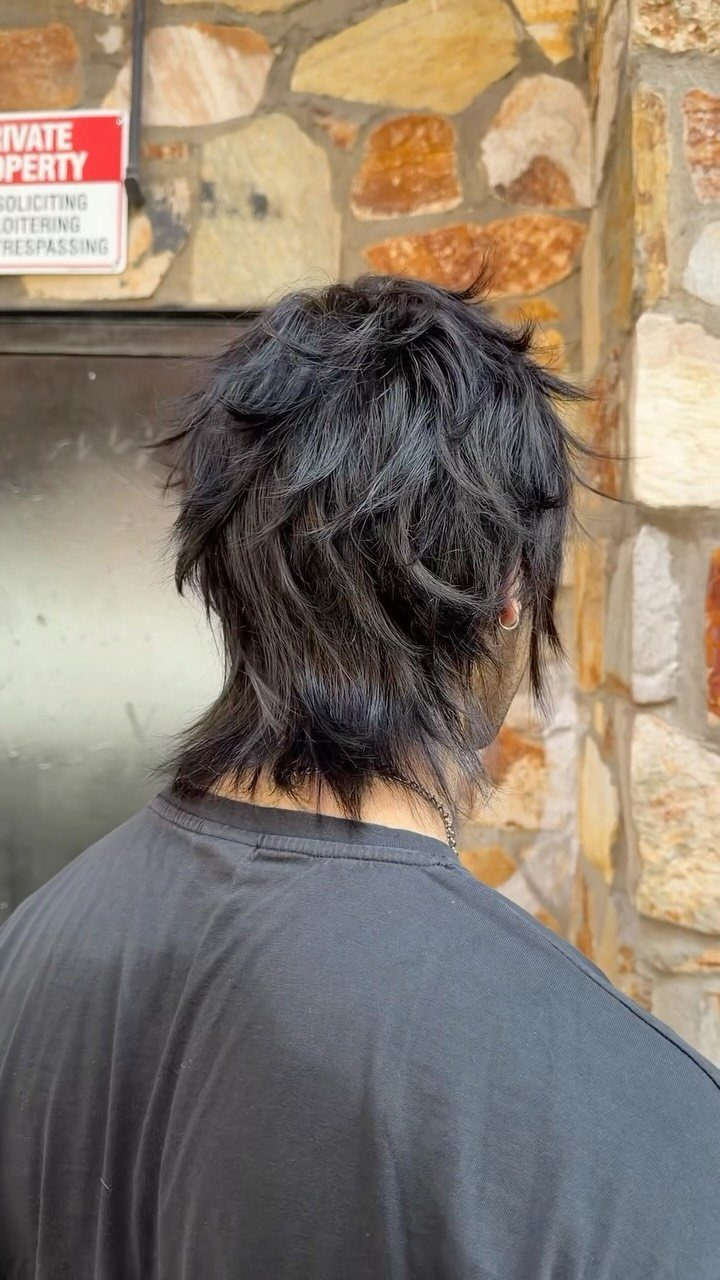 Man with a dark textured wolf cut hairstyle posing against a stone wall.