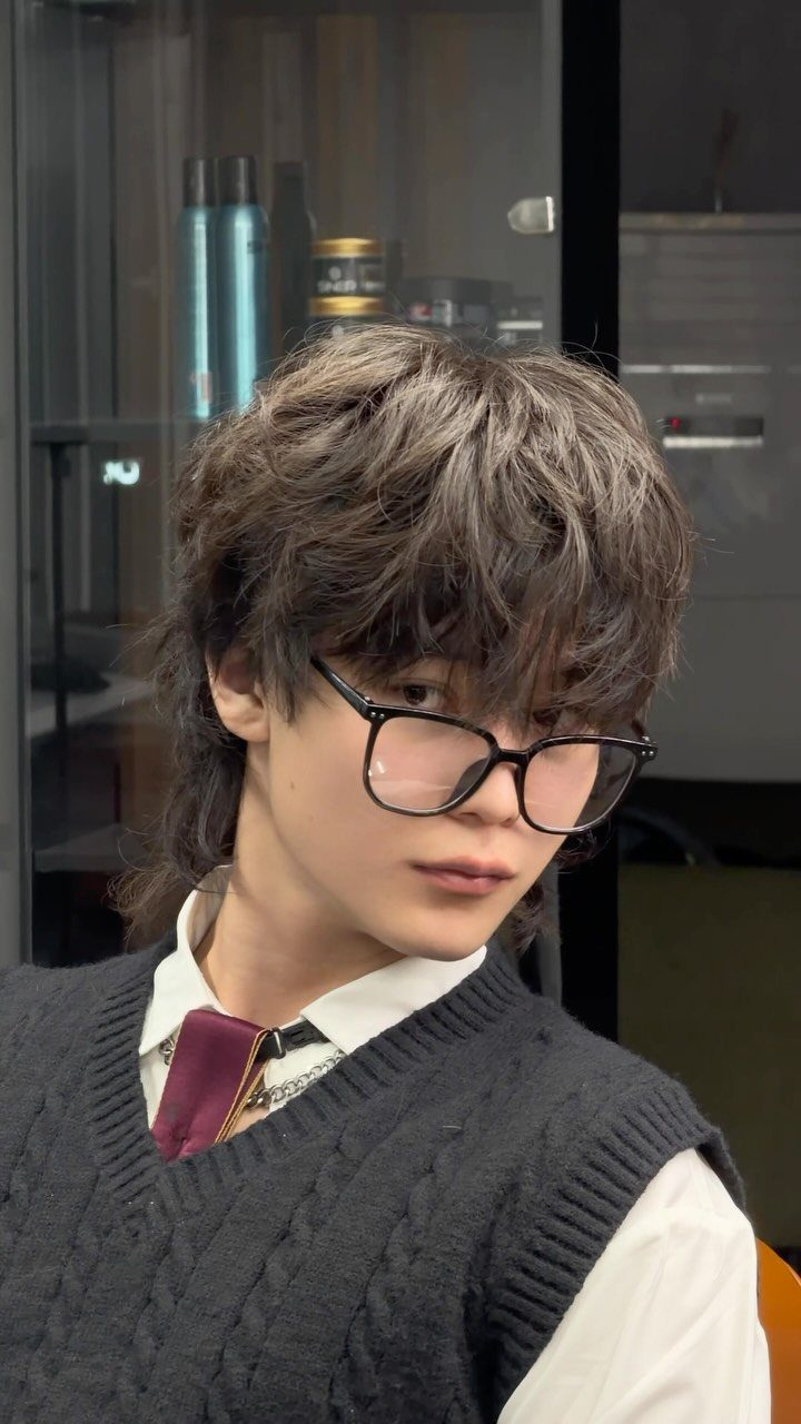 Young man with a brown layered wolf cut wearing glasses and a sweater vest.